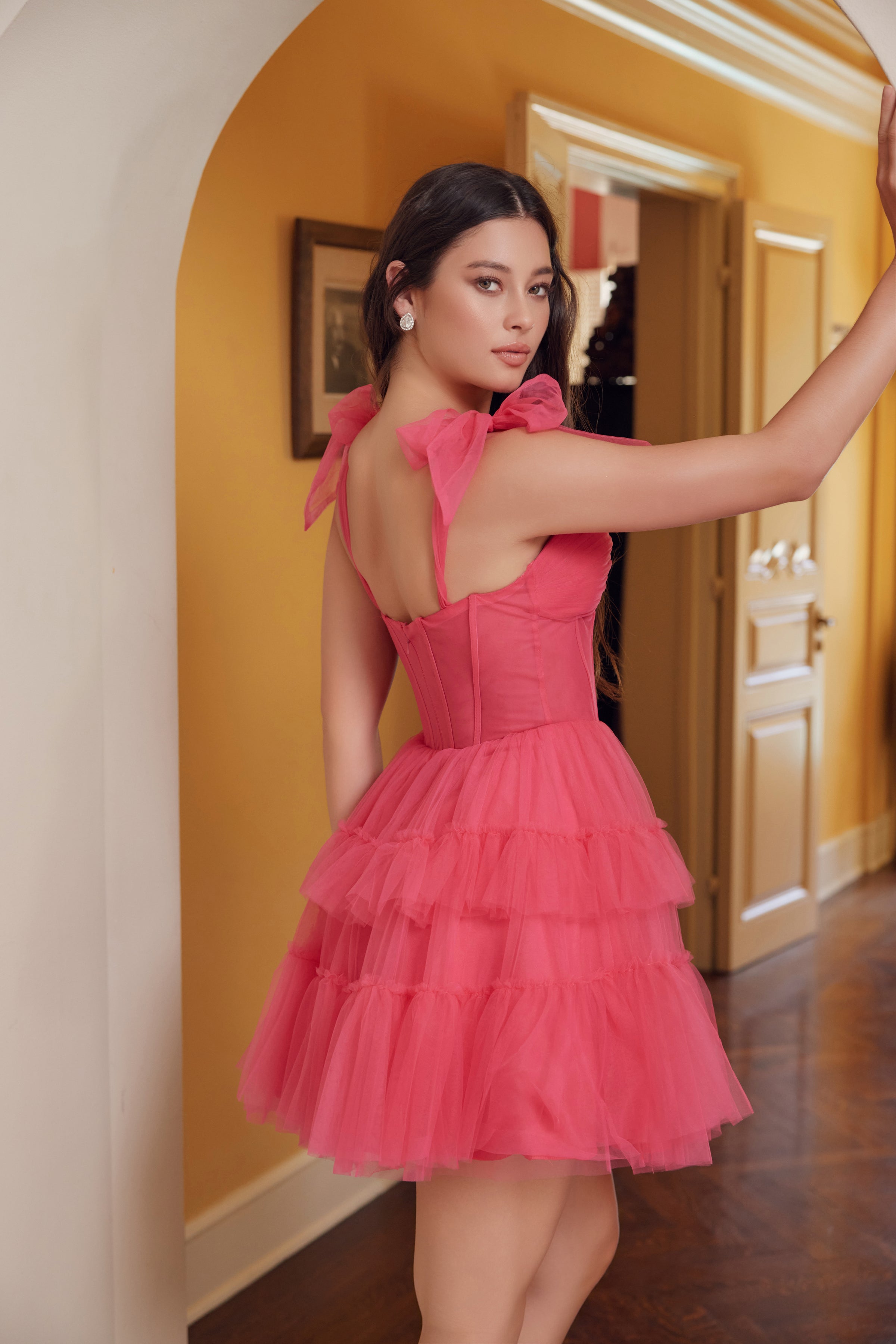 A woman wearing the Shoulder-Tie Short Ruffled A-Line Hoco Dress Y858 stands indoors, showing off its sleeveless tiered pink design with bow straps while looking over her shoulder in a hallway with yellow walls and wooden floors.